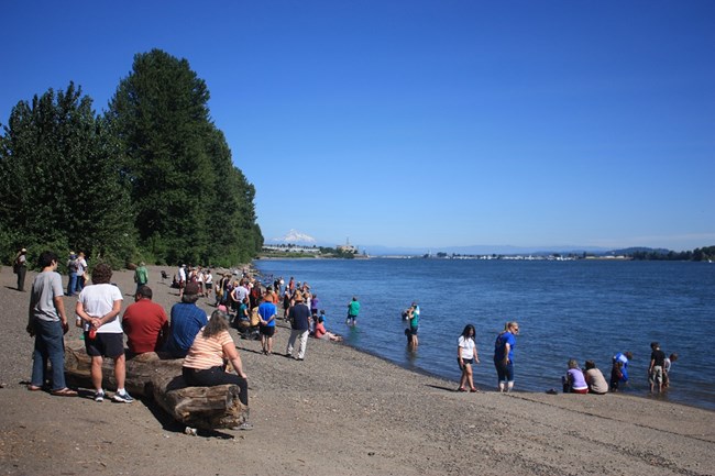 People stand on a beach near the Columbia River on a sunny day. Mount Hood can be seen in the distance.