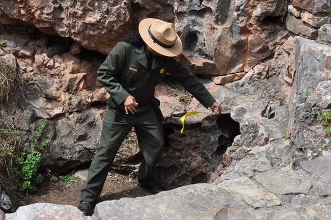 a ranger holding yellow flagging tape in front of a small hole in the rocks, the tape is blowing toward the ranger