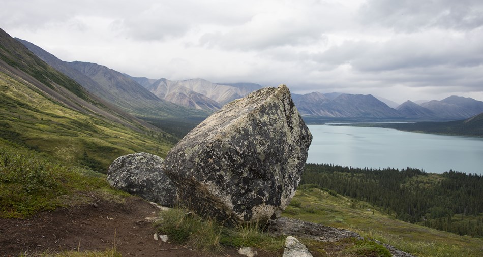 a large boulder balanced on a hillside with mountains and a lake in the background a large boulder balanced on a hillside with mountains and a lake in the background