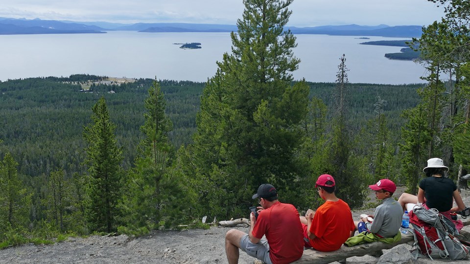 Hikers resting at the top of Elephant Back Mountain Four hikers sit on a rock at the top of the mountain, with a view of the lake in the distance.