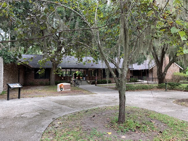 View of the frontside of the Visitor Center blanketed by Live Oak trees with Spanish Moss draped on branches.