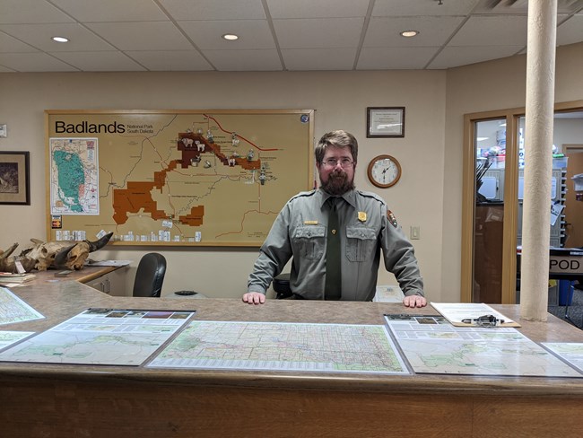 a bearded park ranger stands behind an information desk with a park map in the background.