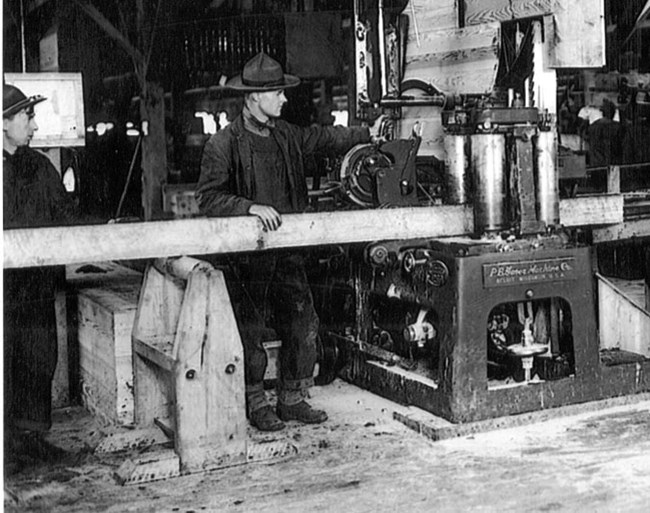 A black and white photo of a man in coveralls feeding a wood board into a saw.