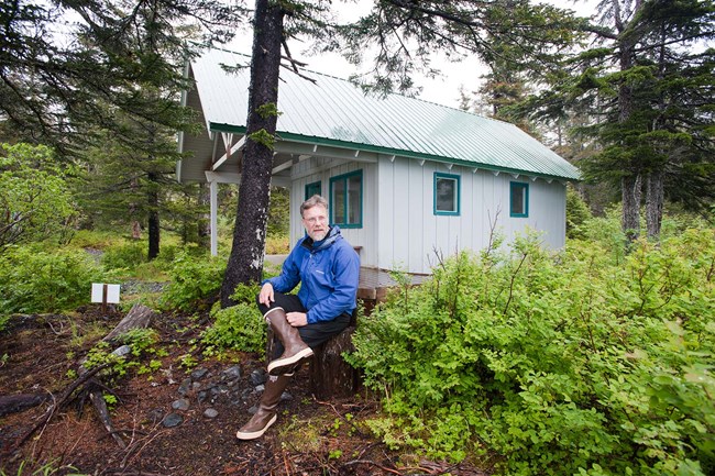 Holgate Public Use Cabin A man sits on a log in the woods, in front of a single room cabin.