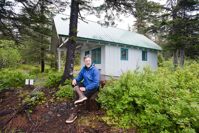 A man sits on a log in the woods, in front of a single room cabin.