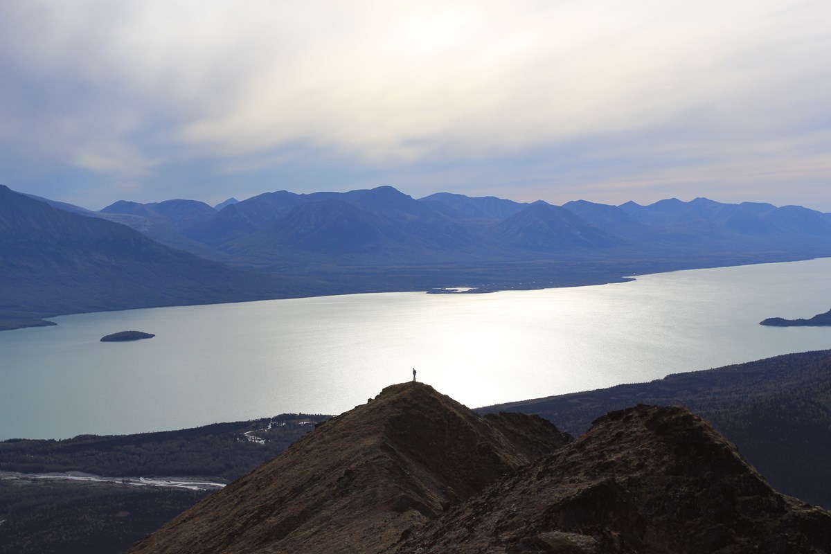 A hiker stands on a rocky peak above Lake Clark