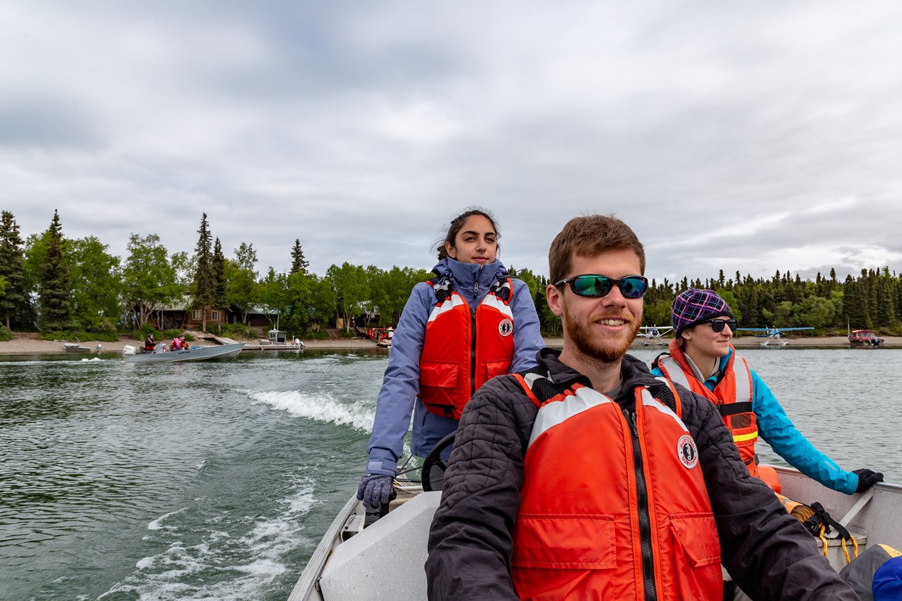 3 people in PFDs are smiling while driving a boat