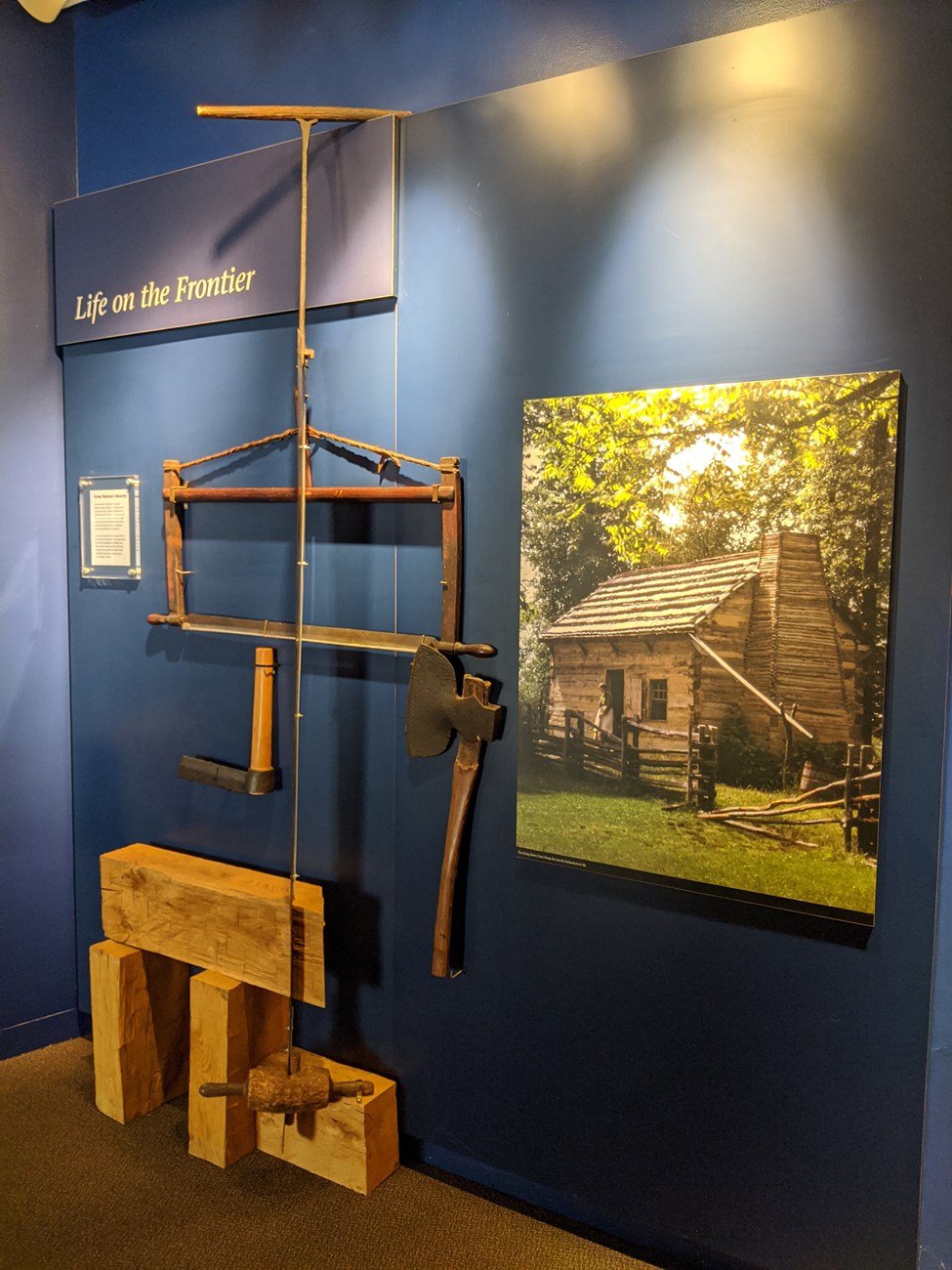 Life on the Frontier museum exhibit Tools used by pioneers hanging on wall alongside a picture of a log cabin