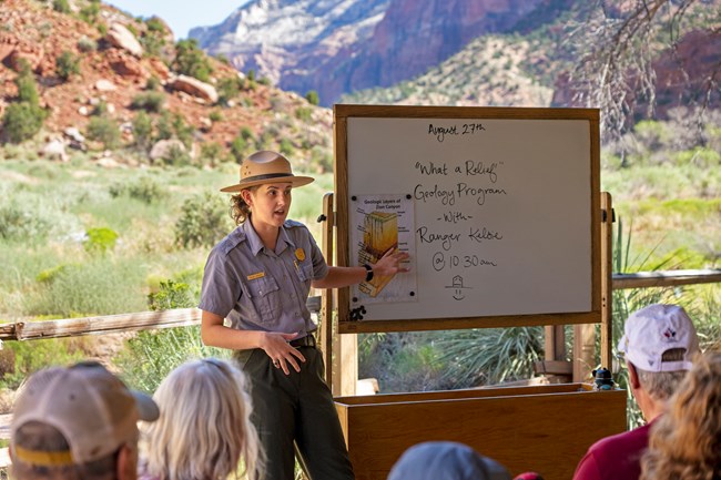 A ranger in a tan flathat stands by a whiteboard in front of seated visitors.