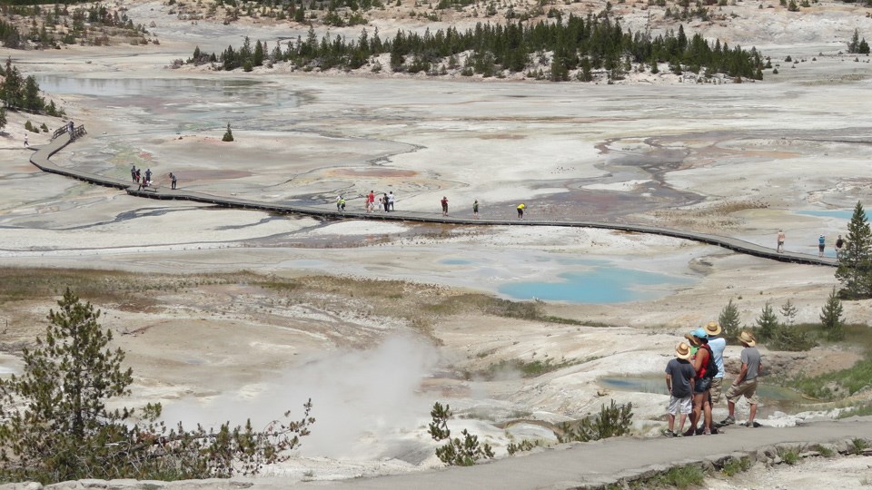 Visitors stand on the paved path looking over the geyser basin, as well as along a boardwalk cutting through the basin.