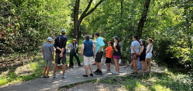 Ranger in a green and grey uniform speaks with a group of visitors outside along a trail.