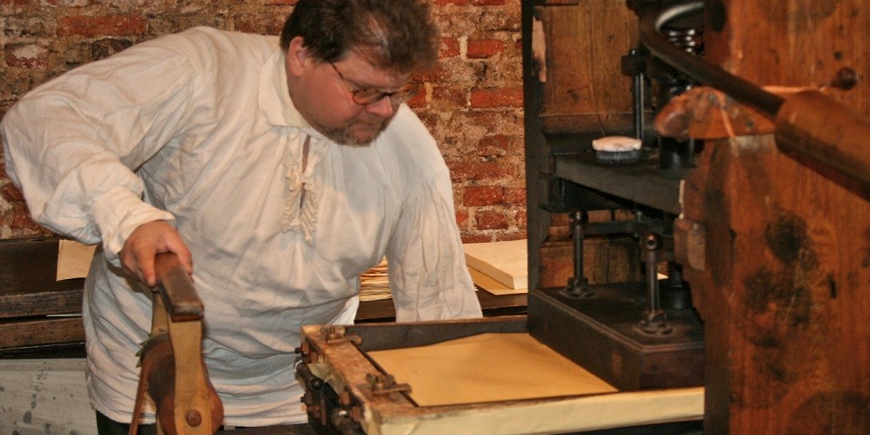 Color photo of man in 18th-century costume demonstrating on a printing press. Color photo of man in 18th-century costume demonstrating on a printing press.