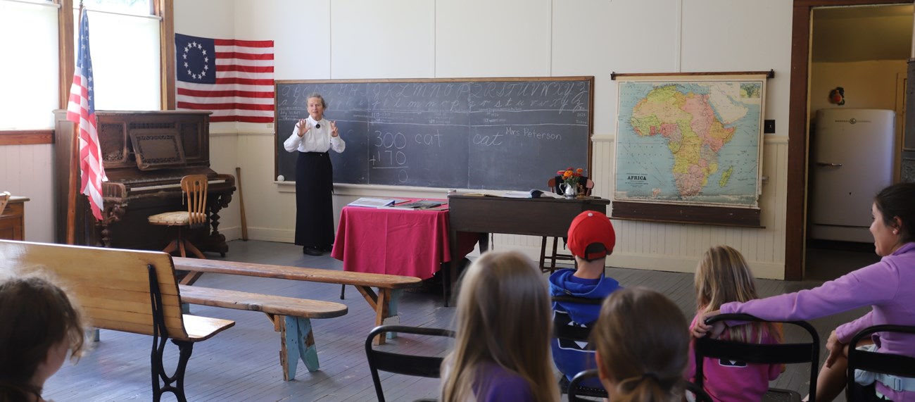 POSchoolHouse-2022 Inside of a historic one-room schoolhouse, surrounded by a piano, chalkboard with the alphabet, and a map of Africa, Mrs. Peterson dressed in a long black skirt and white button-up shirt teaches today's youth.