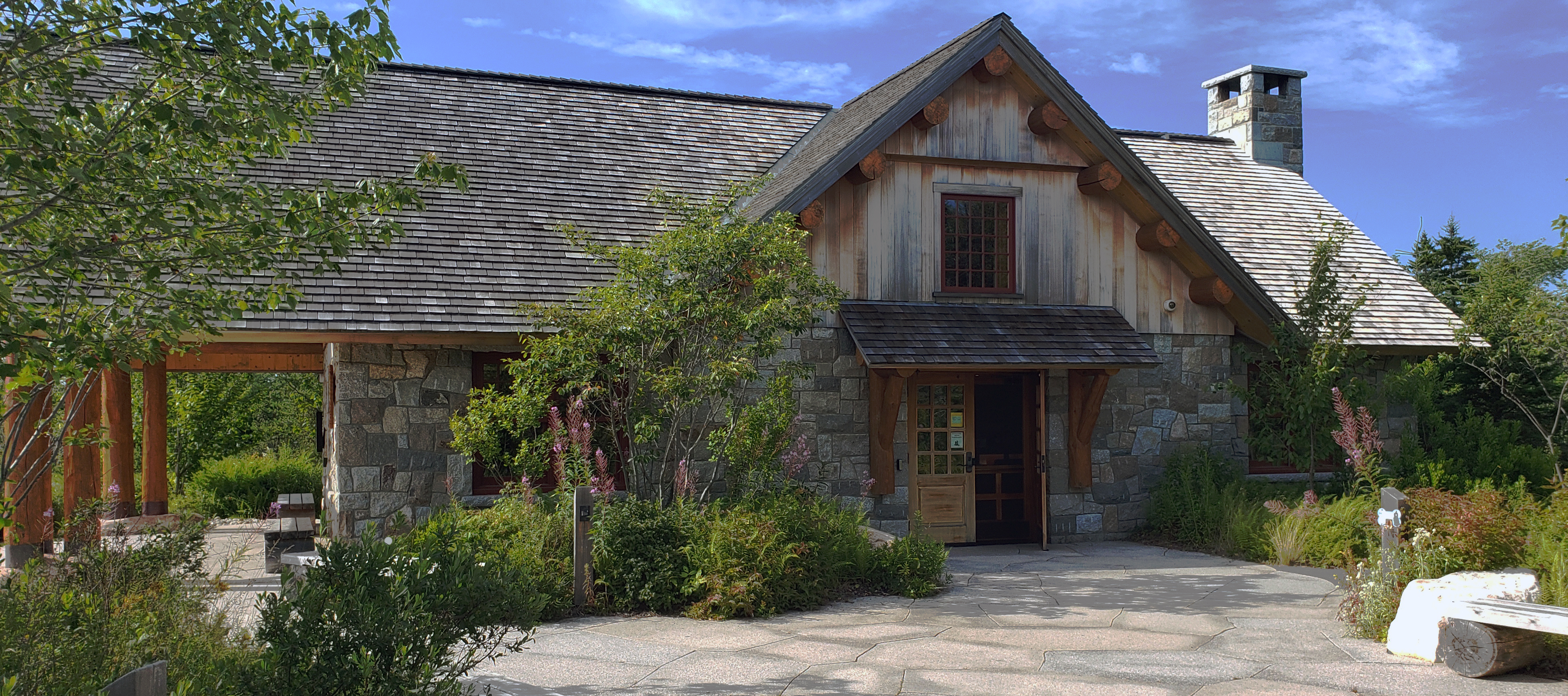 Large, long building with stone walls and chimney, peeled log timbers, and cedar shingles with a stone patio entrance