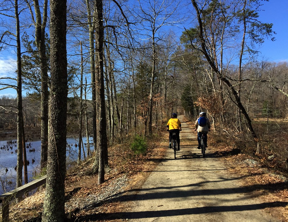 Two bicyclists ride along a path with a pond to the left. Two bicyclists ride along a path with a pond to the left.
