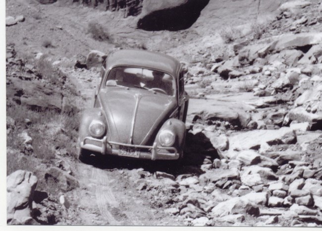 Black and white photo of old VW Bug traversing a rocky unpaved road.