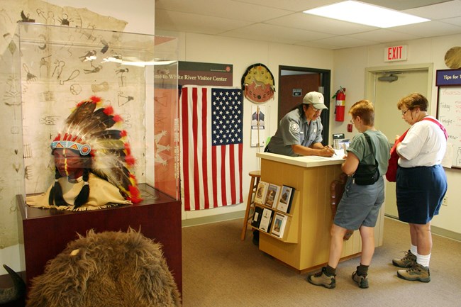 ranger talking to two visitors at an information desk with a headdress display in the foreground