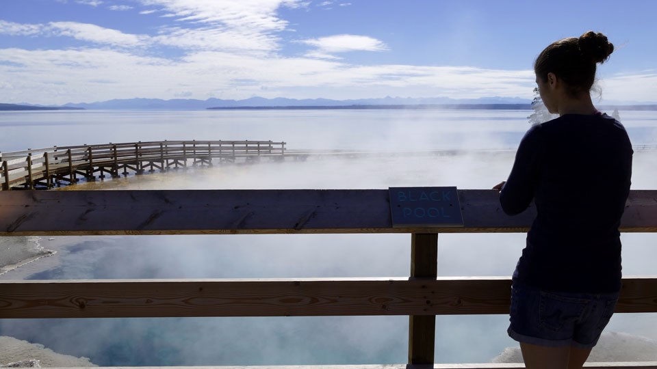 Woman at Black Pool Woman stands by wooden sign that says "Black Pool" and looks at the blue, steaming hydrothermal pool