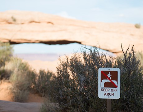 A white warning sign is places in front of a red rock arch. The sign in white with a brown drawing of a person standing on an arch. There is a red slash through it and it says "Keep Off Arch"