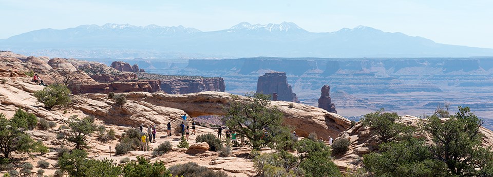 a group of people stand near an arch with distant mountains and canyons in the background