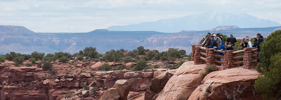 people stand behind wooden fence rails at an overlook. Canyons lie beneath the overlook. people stand behind wooden fence rails at an overlook. Canyons lie beneath the overlook.
