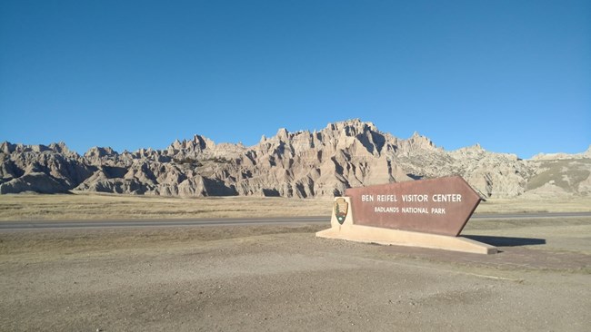 a sign reading "Ben Reifel Visitor Center Badlands National Park" in front of a road and badlands formations