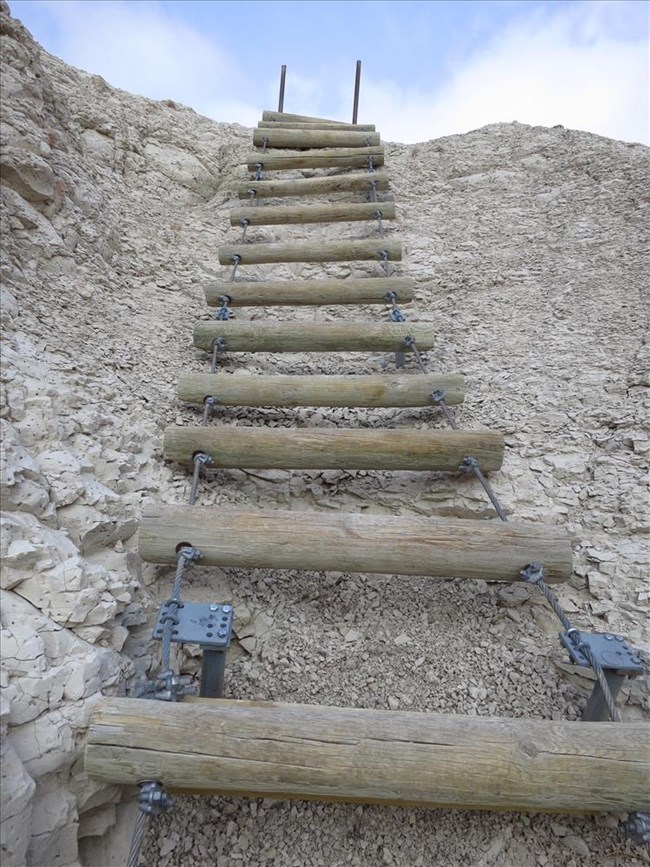 a wooden ladder going up and over a steep formation with blue sky and clouds above