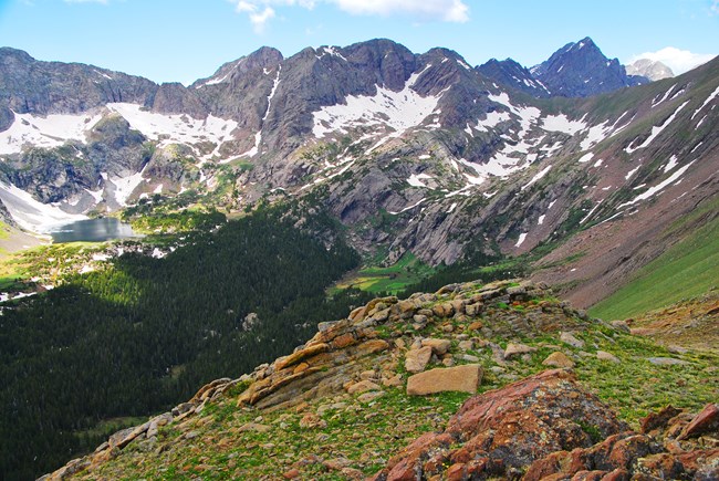 View from green tundra slope into an alpine basin, with a lake at left and snowy peaks above