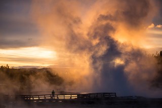 Beautiful reds and golds color a sunset over a boardwalk with a geyser erupting in the background.