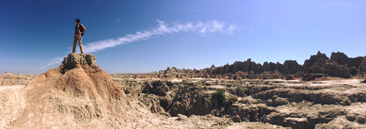 a woman in a polo and khakis stands on top of a badlands formation with buttes and canyon extending out behind her.