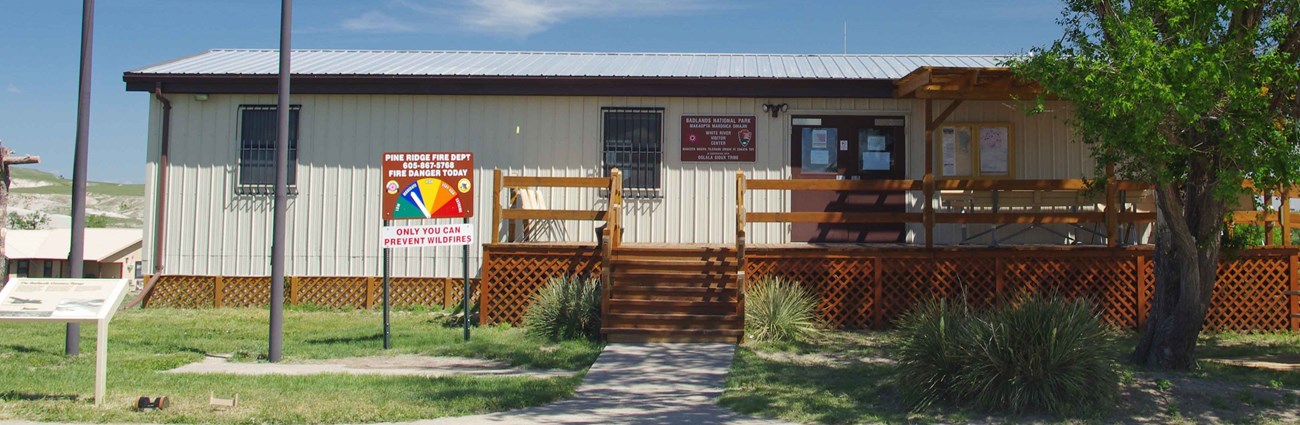 a squat tan building with a brown porch sits beyond green grass and a tree.