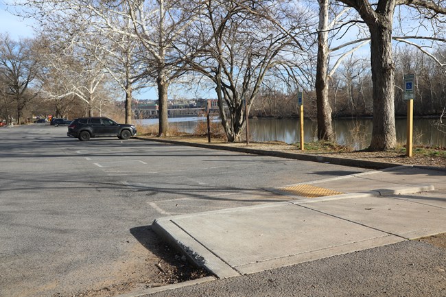 Four empty parking spaces marked with reserved accessible parking signs next to a gradual ramp to a sidewalk along a river lined with trees.