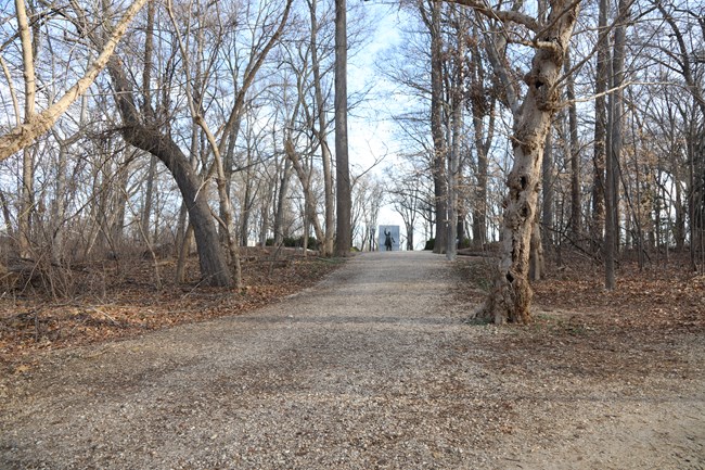 A wide gravel path with trees on either side leads uphill towards a large statue of a man in the distance.