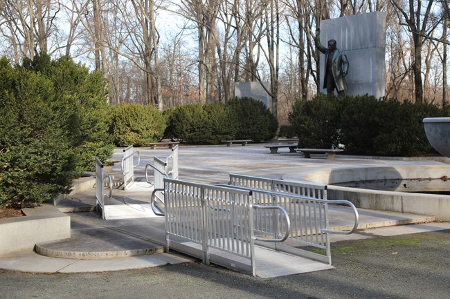 A series of three metal ramps with railings gradually lead up to a paved plaza with hedges, benches, and a large bronze statue of Theodore Roosevelt.