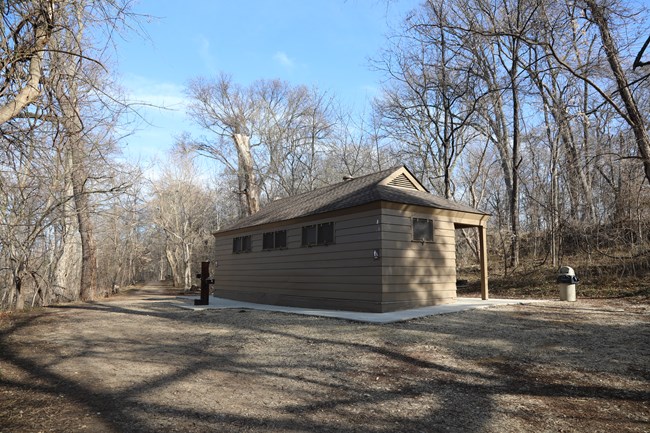 A single-story rectangular brown building with a concrete path perimeter next to a wide gravel path surrounded by trees.