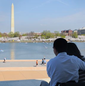 Couple looks off in distance towards Egyptian obelisk reflecting in water surrounded by trees