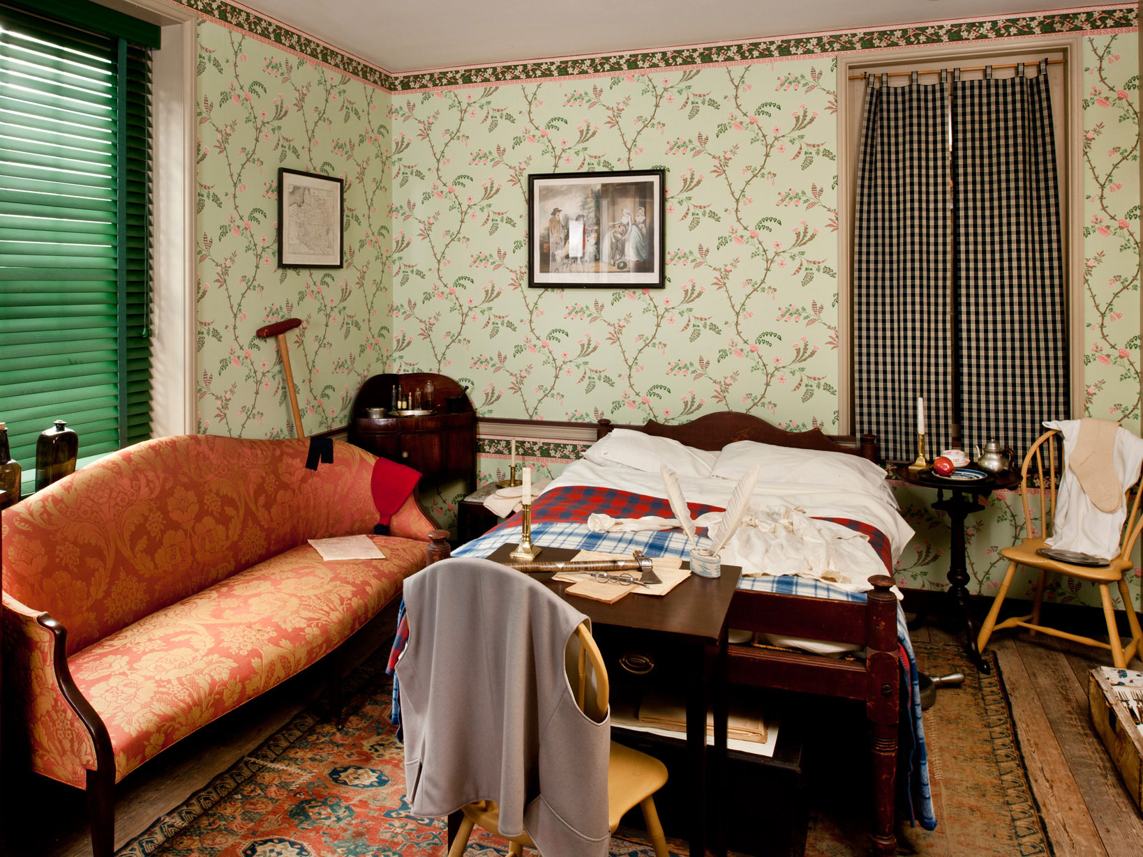Small bedroom with a red couch, red rug, green floral wallpaper, and personal items.