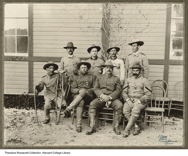 Set of 8 uniformed men (rough riders) sitting in pose for a group photo. 4 men sit in the front row on chairs. The one to the farthest left has crutches. 4 other men stand in uniform behind the first row of sitting men.