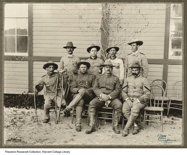 Set of 8 uniformed men (rough riders) sitting in pose for a group photo. 4 men sit in the front row on chairs. The one to the farthest left has crutches. 4 other men stand in uniform behind the first row of sitting men.