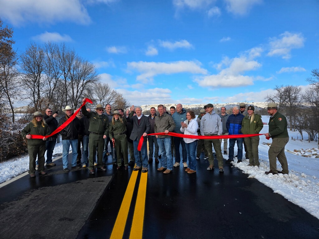 A group of about twenty people stands on a newly paved road in a snowy landscape, gathered for a ribbon-cutting ceremony.