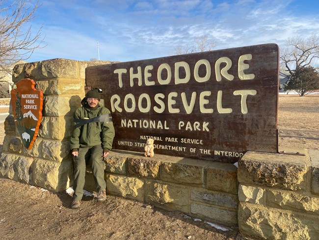 Ranger Peelee sits in front of the park sign with tape around his arms, looking sad. A prairie dog plushie sits next to him.