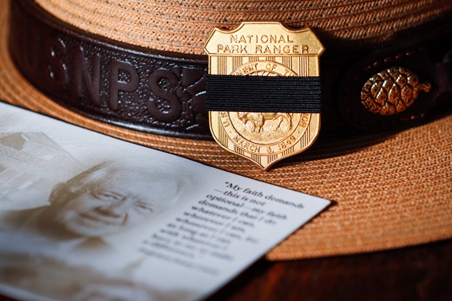 A Park Ranger's flat hat rests on a surface, prominently displaying the National Park Service badge on the front. A black mourning band wraps around the badge. The memorial card for Jimmy Carter sits overlapping the hat reads, "My faith demands—this is no