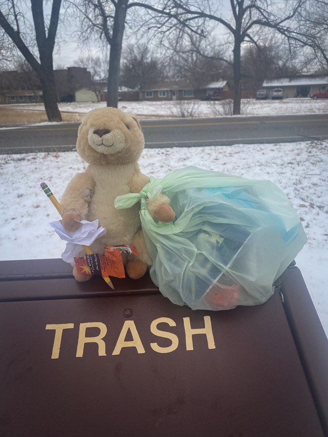 A prairie dog plushie sits on top of a trash bin holding a small plastic bag full of trash and a pencil with some trash poked onto it.