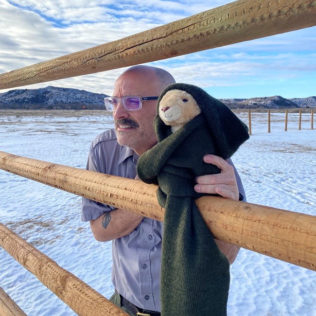 Ranger Peelee and a prairie dog plushie look over a fence into the snowy park. The plushie looks warm with a hat and scarf on, while Ranger Peelee is shivering without a jacket.