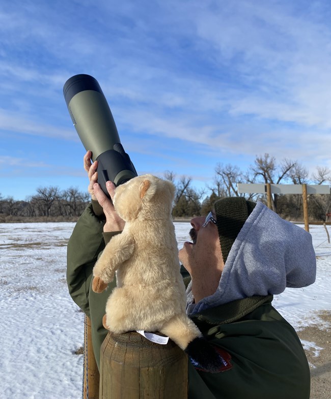 Ranger Peelee holds a spotting scope, pointed up at the sky, in front of a prairie dog plushie.
