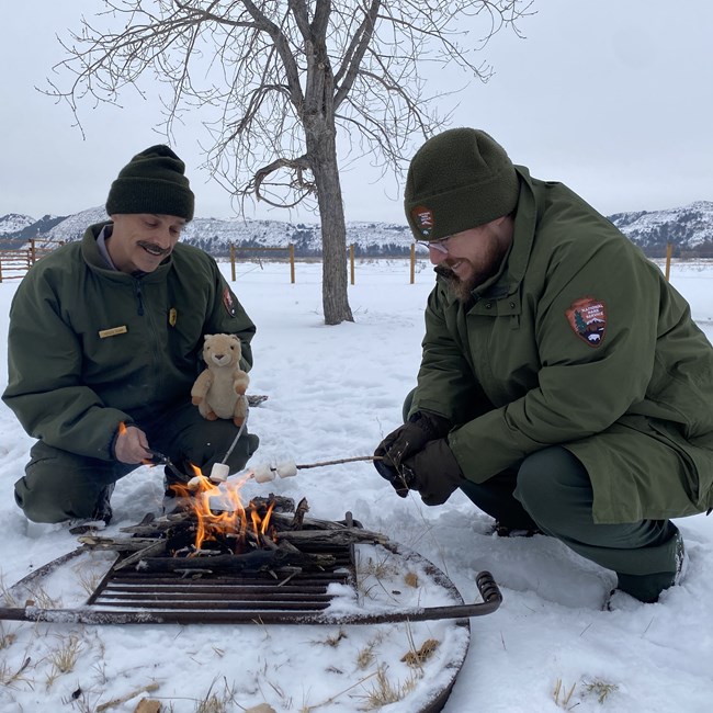 Two male rangers dressed in coats, gloves and hats sit with a prairie dog plushie, roasting marshmallows around a fire in a fire ring.