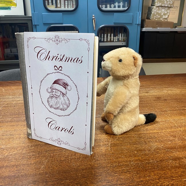 A prairie dog plushie reads from a book entitled "Christmas Carols."