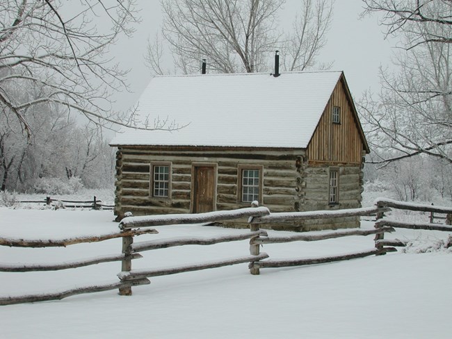 Maltese Cross cabin covered in snow with a wooden fence in the foreground.