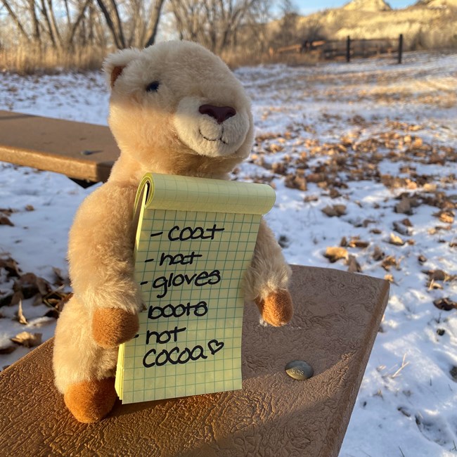 A prairie dog plushie sits on a picnic bench with snow and buttes in the background, holding a written list that says: "coat, hat, gloves, boots, hot cocoa".