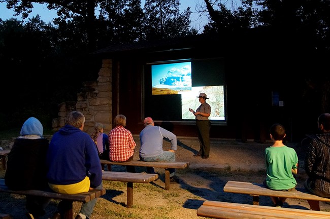 A ranger stands in front of a projection screen while visitors sit on amphitheater benches